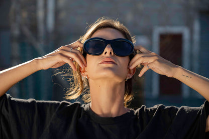 Woman wearing avant-garde party sunglasses with hands framing her face — sculptural oversized cat-eye worn with deliberate editorial confidence, the statement frame that makes every backdrop irrelevant | Sunglass Station