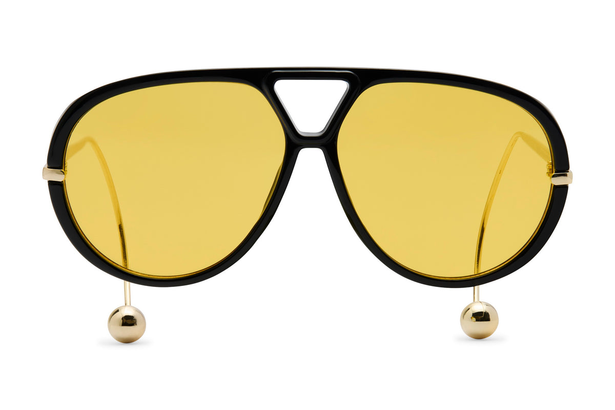 Aviator-style sunglasses with a thick black frame, yellow tinted lenses, and gold-tone metal details including small hanging spheres at the lower corners of the frame.
