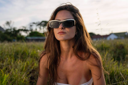 Woman wearing gold and bold party sunglasses outdoors at golden hour — dual-tone parallel frame in sustainable recycled polyester, the statement eyewear that photographs as well as it feels, free delivery UK & Europe | Sunglass Station