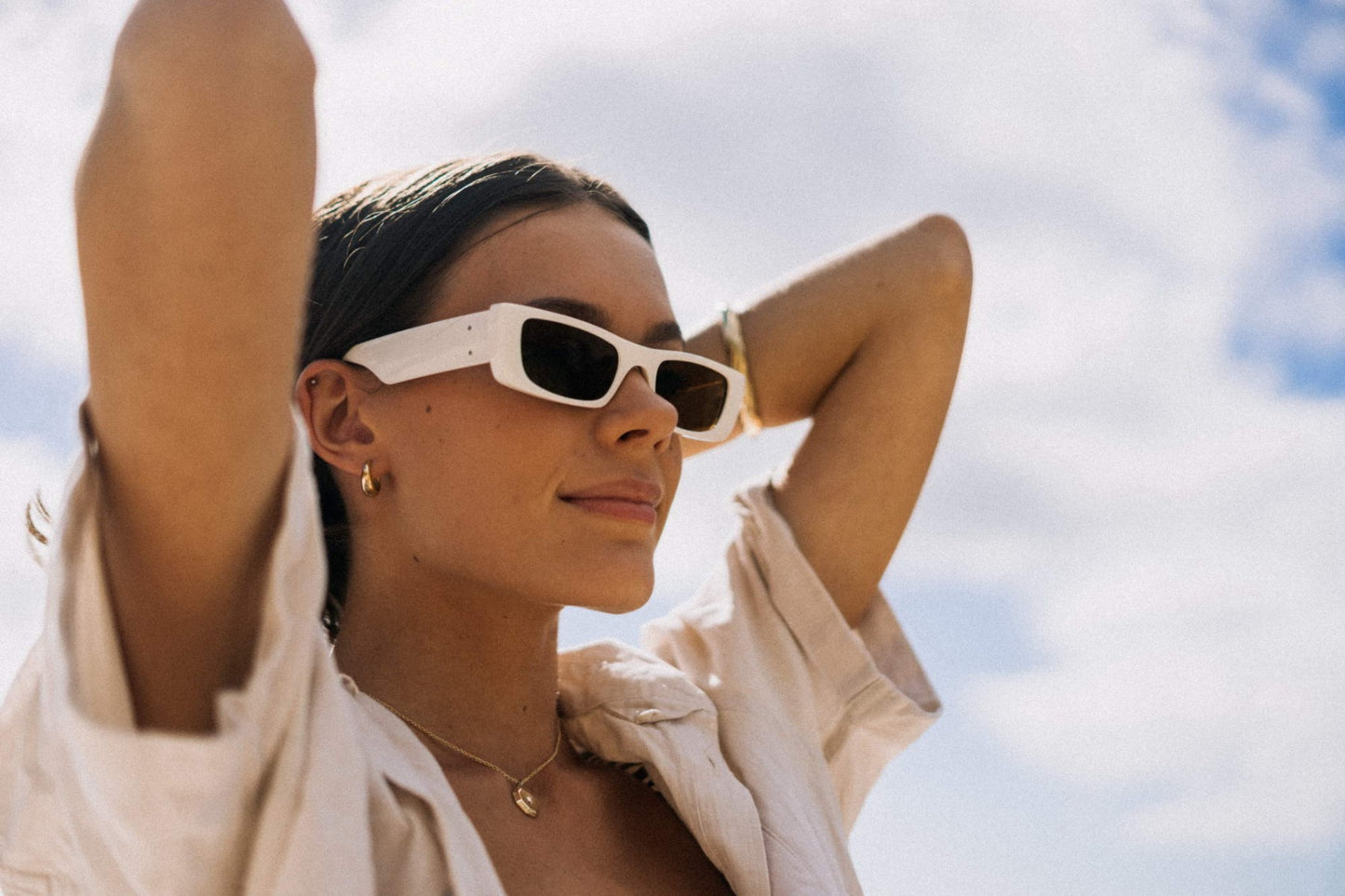 Woman wearing sleek rectangle party sunglasses against a blue sky — slim low-profile polycarbonate frame worn with effortless confidence, the versatile rectangle that moves from city break to festival without changing register | Sunglass Station