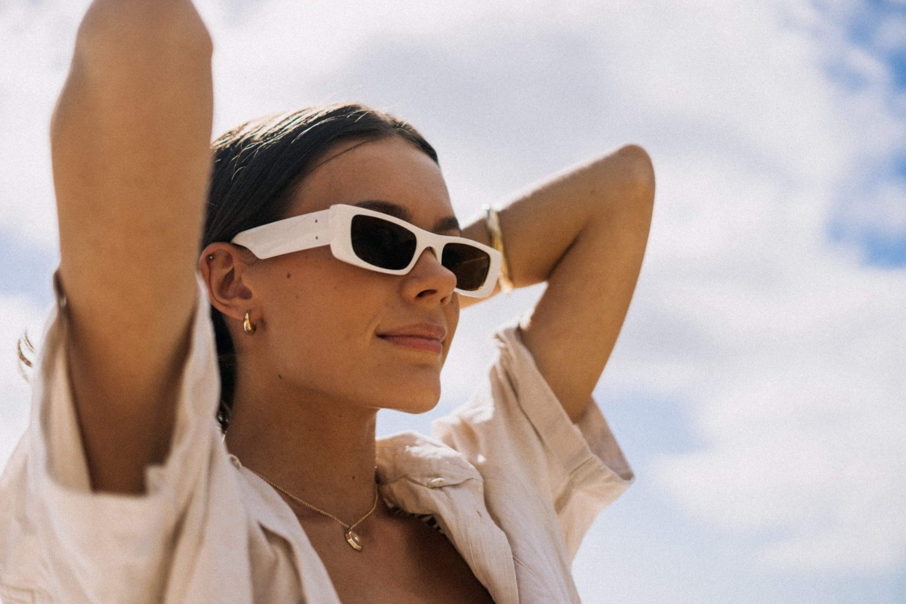 Woman wearing sleek rectangle party sunglasses against a blue sky — slim low-profile polycarbonate frame worn with effortless confidence, the versatile rectangle that moves from city break to festival without changing register | Sunglass Station